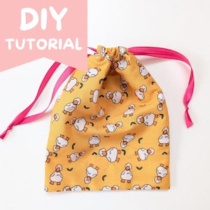Op de afbeelding: Een gele trekkoordtas met een herhaald patroon van witte en oranje cartoon eenden. De tas heeft een roze lint trekkoord en de woorden "DIY TUTORIAL" in het wit op een roze achtergrond.