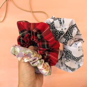 Puede incluir: Una mano sostiene una colecci&oacute;n de coleteros de tela. Uno es de cuadros rojos, otro tiene un estampado de jirafas sobre blanco, y el tercero es floral con acentos lavanda y verdes. Los coleteros presentan una mezcla de texturas y patrones.