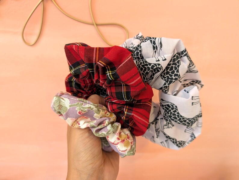 Puede incluir: Una mano sostiene una colecci&oacute;n de coleteros de tela. Uno es de cuadros rojos, otro tiene un estampado de jirafas sobre blanco, y el tercero es floral con acentos lavanda y verdes. Los coleteros presentan una mezcla de texturas y patrones.