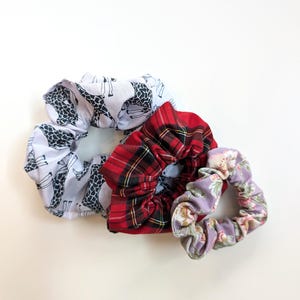 Puede incluir: Tres coleteros de tela con diferentes estampados. Uno es blanco con siluetas de jirafas negras, otro es de cuadros rojos y el tercero tiene un estampado floral con flores rosas y moradas sobre un fondo lavanda. Estos accesorios para el cabello son ideales para a&ntilde;adir un toque de estilo.