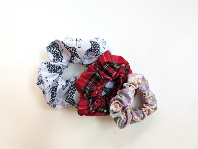 Puede incluir: Tres coleteros de tela con diferentes estampados. Uno es blanco con siluetas de jirafas negras, otro es de cuadros rojos y el tercero tiene un estampado floral con flores rosas y moradas sobre un fondo lavanda. Estos accesorios para el cabello son ideales para a&ntilde;adir un toque de estilo.