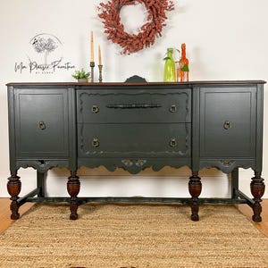 May include: A dark green vintage buffet with a wood top and carved details. The buffet has four legs and two doors on each side. It is sitting on a natural fiber rug in front of a white wall. The buffet is decorated with a wreath and candles.