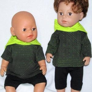 Könnte beinhalten: Zwei Puppen in passenden Outfits. Die Outfits sind schwarz mit einem grünen Schaltkreis-Muster und einer neon-grünen Kapuze. Die Puppen sitzen auf einem weißen Hintergrund.