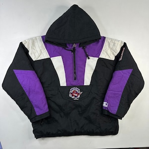 Puede incluir: Una chaqueta con capucha negra, morada y blanca con cierre de cremallera. La chaqueta tiene un gran logotipo de "Raptors" en la parte delantera con un dinosaurio rojo como mascota. La chaqueta tiene un logotipo de Starter en la manga derecha.