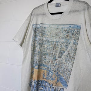 Puede incluir: Camiseta blanquecina con una ilustraci&oacute;n detallada de una escena de un partido de baloncesto. El gr&aacute;fico incluye las palabras "North Carolina" en azul y la frase "We Tar" en la esquina superior derecha. La camiseta est&aacute; colgada en una percha negra.
