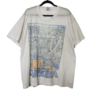 Puede incluir: Camiseta gris claro con una escena de cancha de baloncesto y una multitud de espectadores. Las palabras "North Carolina" est&aacute;n impresas en azul y gris en la parte inferior. La camiseta tiene mangas cortas y cuello redondo.