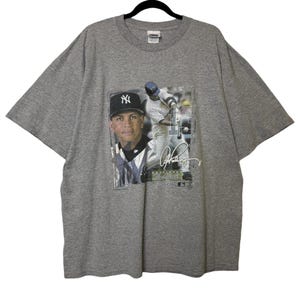 May include: A gray t-shirt featuring a graphic of a baseball player in a New York Yankees uniform. The graphic includes a portrait of the player, his signature, and the team logo. The shirt is made of a soft material.