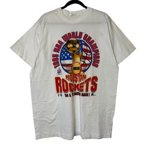 May include: White t-shirt featuring a graphic commemorating the 1995 NBA World Champions, the Houston Rockets. The design includes a trophy, basketball, and the American flag, with the text "Houston Rockets" in red.