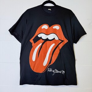 以下が含まれることがあります： オレンジと白のローリング・ストーンズの舌と唇のロゴが特徴の黒いTシャツ。デザインには、白いスクリプトで「Rolling Stones '89」というテキストが含まれています。シャツは黒いハンガーにかかっています。