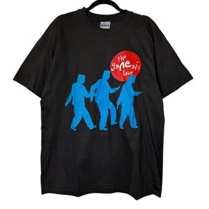 May include: Black t-shirt featuring a blue graphic of three figures walking, with a red circle and the text "the genesis tour". The shirt has a crew neck and short sleeves. The shirt is made of a soft material.