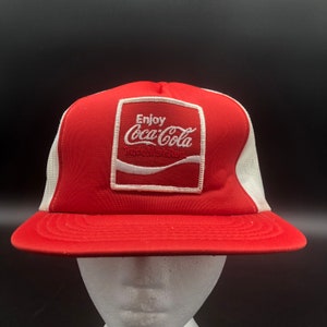May include: Red and white trucker hat with a white Coca-Cola logo and the text "Enjoy Coca-Cola Trade Mark Reg."
