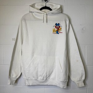 Puede incluir: Sudadera con capucha blanca con bolsillo delantero y cord&oacute;n. La sudadera presenta un gr&aacute;fico colorido de Mickey Mouse con el texto "Minnie California". La sudadera est&aacute; colgada de una percha negra.