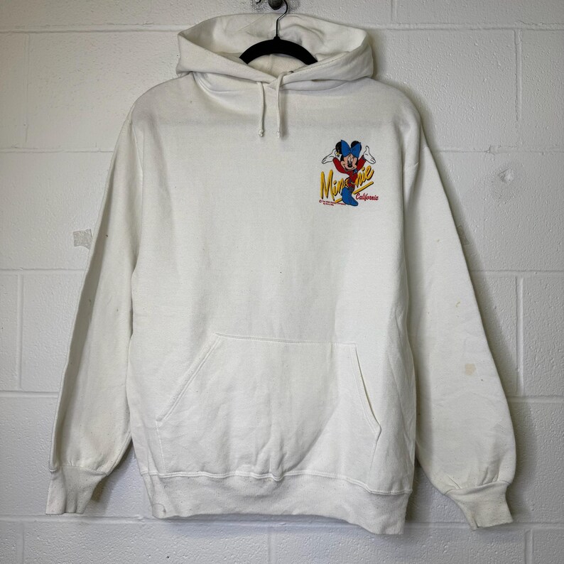 Puede incluir: Sudadera con capucha blanca con bolsillo delantero y cord&oacute;n. La sudadera presenta un gr&aacute;fico colorido de Mickey Mouse con el texto "Minnie California". La sudadera est&aacute; colgada de una percha negra.