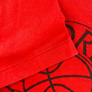 May include: Close-up of a red t-shirt with a black basketball graphic. The shirt has a ribbed texture and a folded hem.