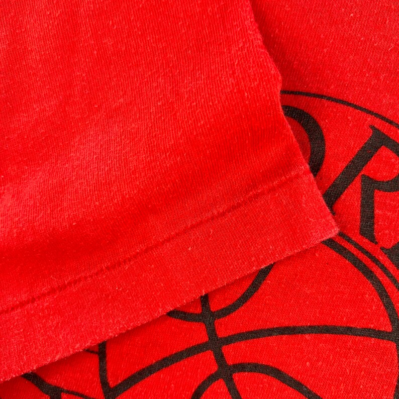 May include: Close-up of a red t-shirt with a black basketball graphic. The shirt has a ribbed texture and a folded hem.
