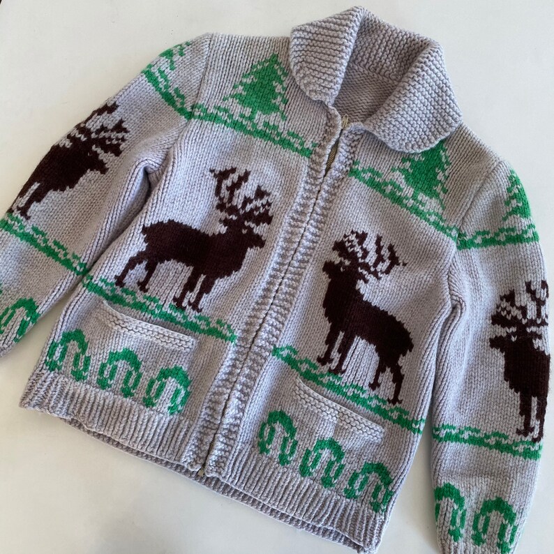 May include: A gray knitted cardigan with a zipper closure and a pattern of brown reindeer and green trees. The cardigan has two pockets.