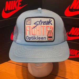 May include: A light blue trucker hat with a black and white patch that says "Streak Fighter" and "Optikleen" with a pink and blue bottle graphic.