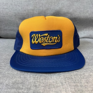May include: A trucker hat with a yellow crown and a blue brim. The hat features a blue mesh back and a rectangular patch with the embroidered text "Weston's" in gold lettering.
