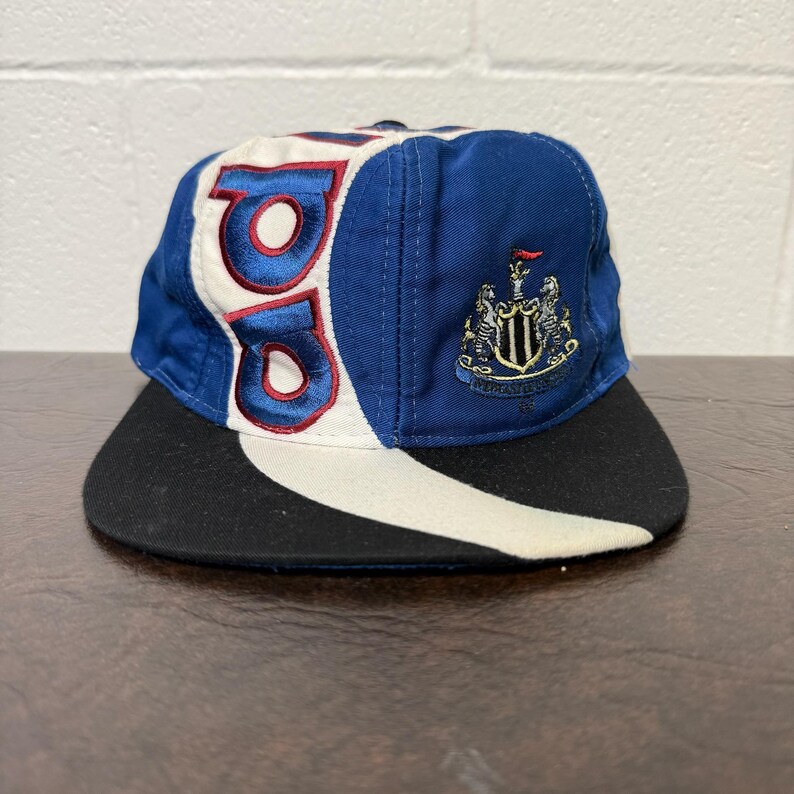 May include: A blue, white, and black baseball cap with the letters "BB" in red embroidery. The cap features a team crest on the side and a black brim. The hat is made of fabric.