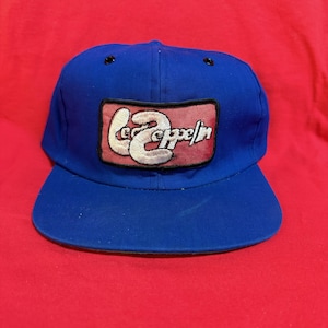 Puede incluir: Gorra de béisbol azul con visera curva. La gorra presenta un parche rectangular con el texto "Led Zeppelin" en escritura blanca sobre un fondo rosa. Diseño clásico de seis paneles.