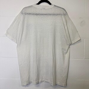 Puede incluir: Una camiseta blanca de manga corta colgada de una percha negra. La camiseta est&aacute; hecha de un material fino y ligeramente transparente. La parte trasera de la camiseta es visible, mostrando el escote y la forma general de la prenda.