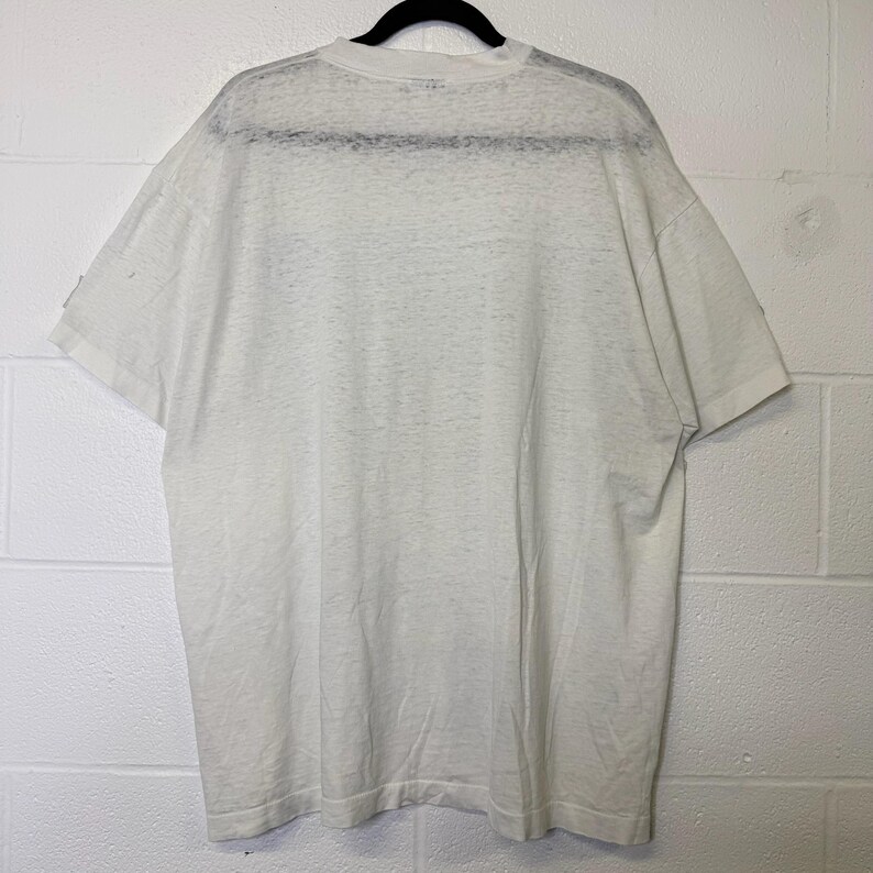 Puede incluir: Una camiseta blanca de manga corta colgada de una percha negra. La camiseta est&aacute; hecha de un material fino y ligeramente transparente. La parte trasera de la camiseta es visible, mostrando el escote y la forma general de la prenda.