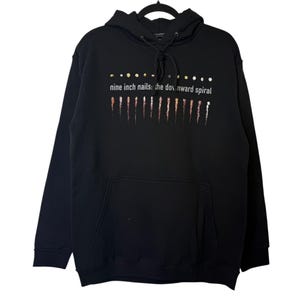 Puede incluir: Sudadera con capucha negra con bolsillo delantero y cordón. La parte delantera de la sudadera presenta el texto "nine inch nails: the downward spiral" sobre una serie de líneas verticales de colores.