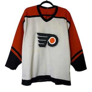 May include: A vintage hockey jersey with a white body, orange sleeves, and black accents. The jersey features a V-neck and the Philadelphia Flyers logo on the chest. The logo is a black and white winged emblem with an orange circle.