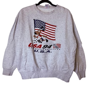 May include: A light gray sweatshirt featuring a cartoon dog holding a soccer ball and an American flag. The graphic includes the text "USA 94 U.S.A." in red and blue. The sweatshirt has a crew neck and ribbed cuffs and hem.