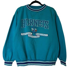 May include: Teal-colored Hornets sweatshirt with the team name in large, white, outlined letters. Below the name is a logo of a cartoon hornet and the words "National Basketball Association". The collar and cuffs have blue and white stripes.