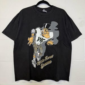May include: Black t-shirt with a cartoon image of a man in a top hat and football uniform. The graphic includes the letters "WF" and the text "White Forest Demons". The t-shirt is displayed on a black hanger.