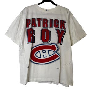 May include: White t-shirt with the name "PATRICK ROY" in red block letters with a blue outline. Below the name is the Montreal Canadiens logo in red, white, and blue. The shirt has short sleeves and a crew neck.