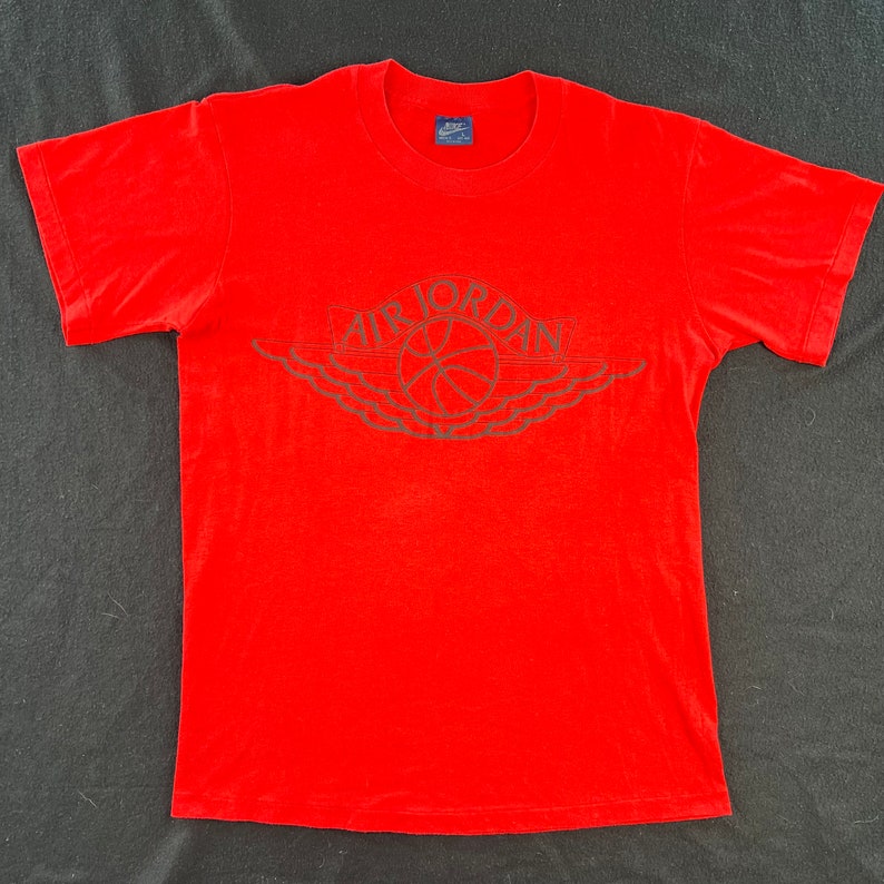 May include: Red t-shirt with a black and white Air Jordan logo. The logo features a basketball and wings.
