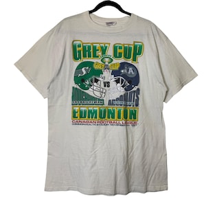 May include: Off-white t-shirt featuring a graphic for the Grey Cup, a Canadian football championship. The design includes two football helmets, one green and one blue, with the text "Grey Cup" and "Edmonton". The shirt also has the text "Canadian Football League, Commonwealth Stadium November 1997."