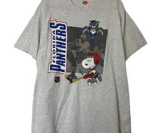 May include: A gray t-shirt featuring the Florida Panthers logo and a Snoopy cartoon character playing hockey. The shirt has a crew neck and short sleeves. The graphic includes the team's logo and a vintage hockey scene.