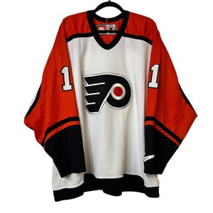 May include: A white, orange, and black hockey jersey with the number 1 on the sleeves. The jersey features the Philadelphia Flyers logo on the front and a Nike logo. The jersey is hanging on a black hanger.