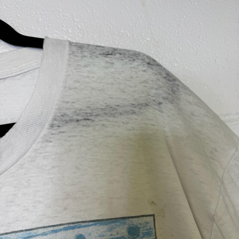 Puede incluir: Primer plano de una camiseta blanca con cuello redondo. La camiseta tiene un aspecto descolorido, ligeramente transparente, con sutiles sombras grises en la zona de los hombros. La tela parece suave y ligera.