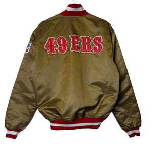 May include: A gold satin bomber jacket with red and white trim. The back of the jacket features the text "49 ERS" in red, outlined in white. An NFL logo patch is visible on the left sleeve.