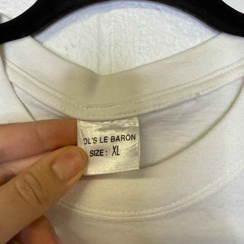 May include: White t-shirt with a tag that reads "DL'S LE BARON SIZE: XL". The tag is rectangular and has black text. The shirt is being held by a person.