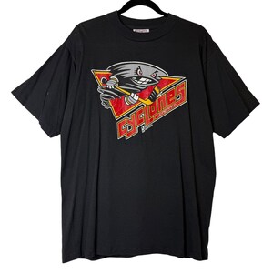 May include: Black t-shirt featuring a red, white, and gray cartoon cyclone mascot holding a hockey stick. The word "CYLONES" is printed in red letters on a red triangle. The shirt is hanging on a black hanger.