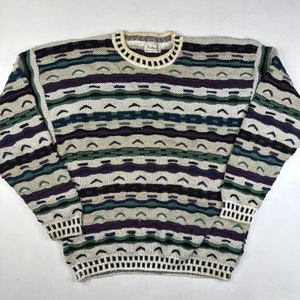 Puede incluir: Un suéter multicolor con un patrón geométrico. El suéter está hecho de una tela suave y tejida y tiene cuello redondo. El suéter es blanco, verde, morado y azul.