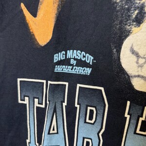 May include: Close-up of a navy blue t-shirt with the text "TAR" in a gradient blue and white font. The shirt also displays the text "BIG MASCOT" above the word "TAR", with the brand name "WAULDRON SPORTS" below it. An orange and white graphic is visible.