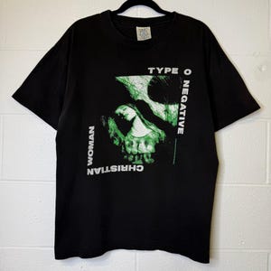 May include: Black t-shirt featuring a green and white graphic design with the text "TYPE O NEGATIVE" and "CHRISTIAN WOMAN". The design includes an image of a woman's face. The shirt is hanging on a black hanger.