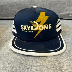 May include: Navy blue trucker hat with white stripes and a white brim. The hat features the text "SKY DOME" in yellow with a lightning bolt design. The text "TORONTO, CANADA" is below the logo.