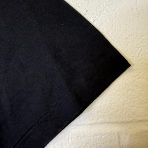 May include: Close-up of a black t-shirt corner. The fabric appears to be a soft knit material. The image is set against a white, textured background.