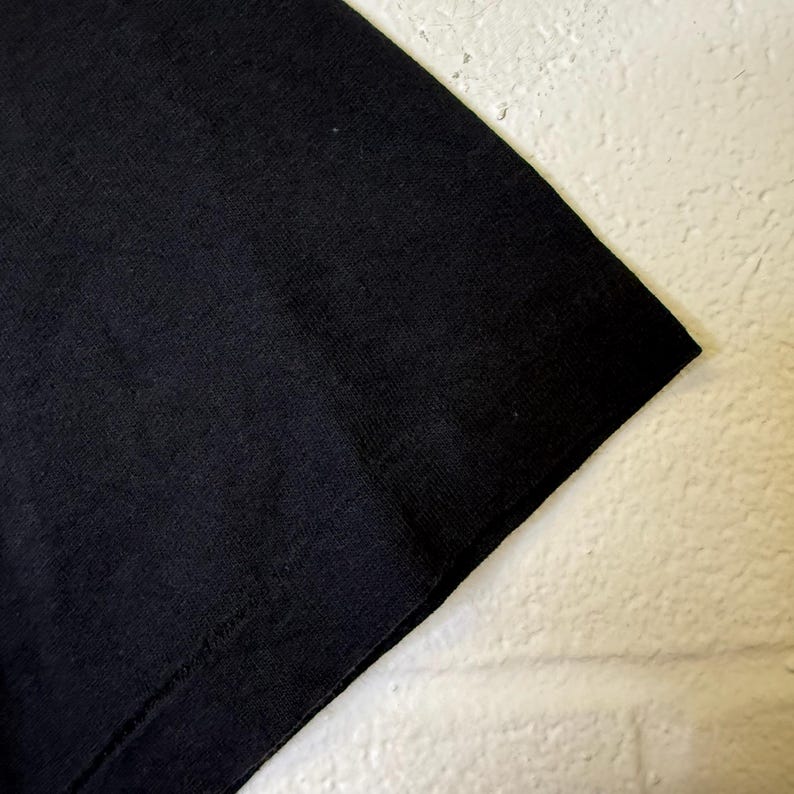 May include: Close-up of a black t-shirt corner. The fabric appears to be a soft knit material. The image is set against a white, textured background.