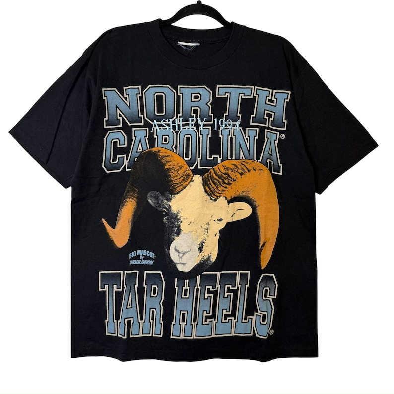 May include: Black t-shirt with a ram graphic and the text "NORTH CAROLINA" and "TAR HEELS". The ram has tan horns and a cream-colored face. The text is in a light blue and white colour scheme.