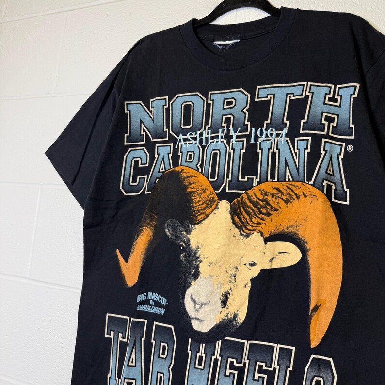 May include: Black t-shirt with a ram graphic, orange horns, and the text "NORTH CAROLINA" in blue and white. The shirt also features "TAR HEELS" and "ASHLEY 1994".