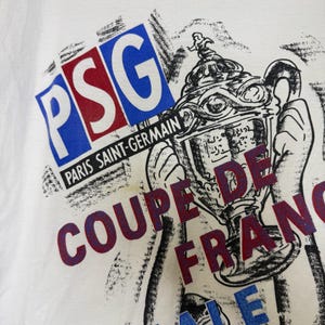 May include: White t-shirt featuring a graphic design with the letters "PSG" in blue and red, and the words "Paris Saint-Germain" and "Coupe de France". The design includes a trophy illustration.