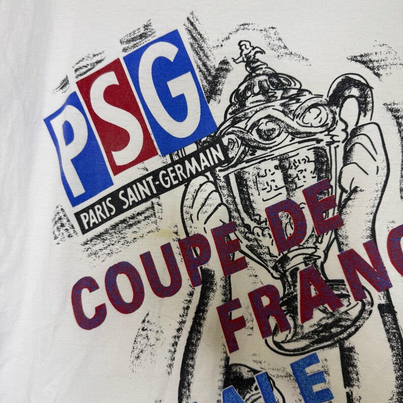 May include: White t-shirt featuring a graphic design with the letters "PSG" in blue and red, and the words "Paris Saint-Germain" and "Coupe de France". The design includes a trophy illustration.